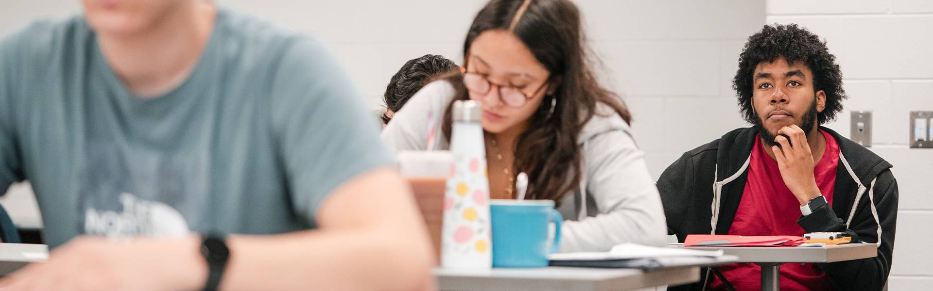 Three students sitting at tables in a classroom looking towards the front of the classroom