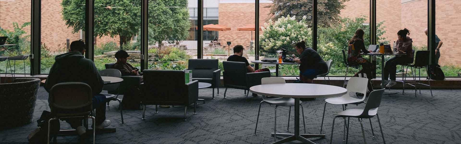 Students studying at different tables in a common area in front of a wall of windows