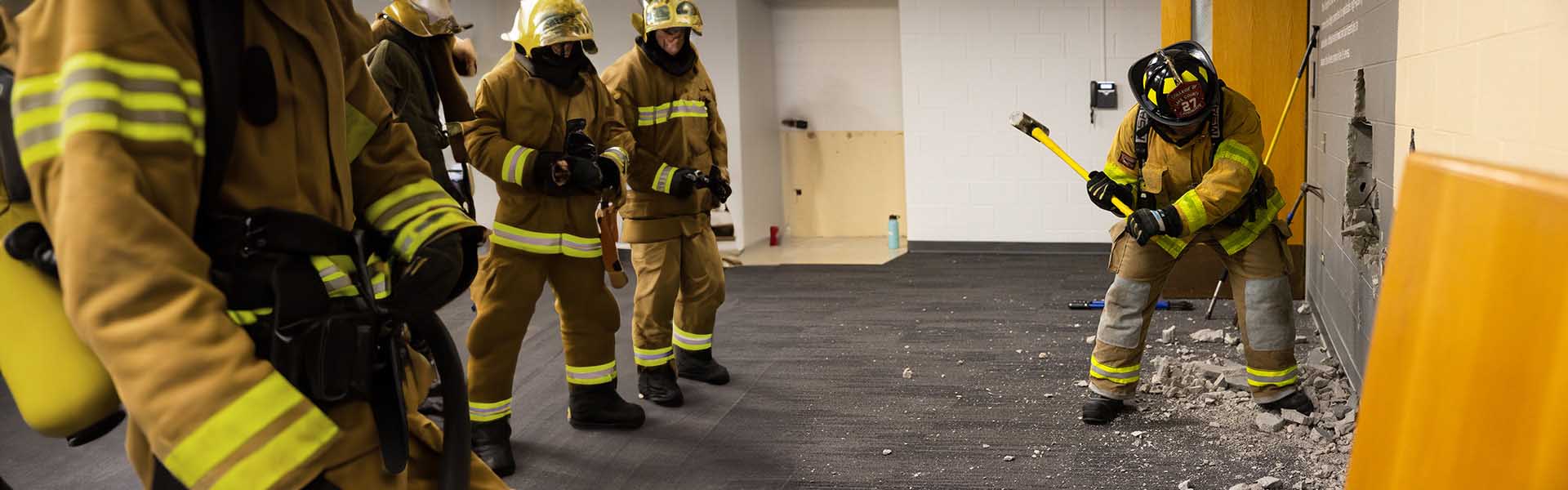 Fire Science Technology student wearing safety gear hitting a wall with a hammer while conducting drill in class