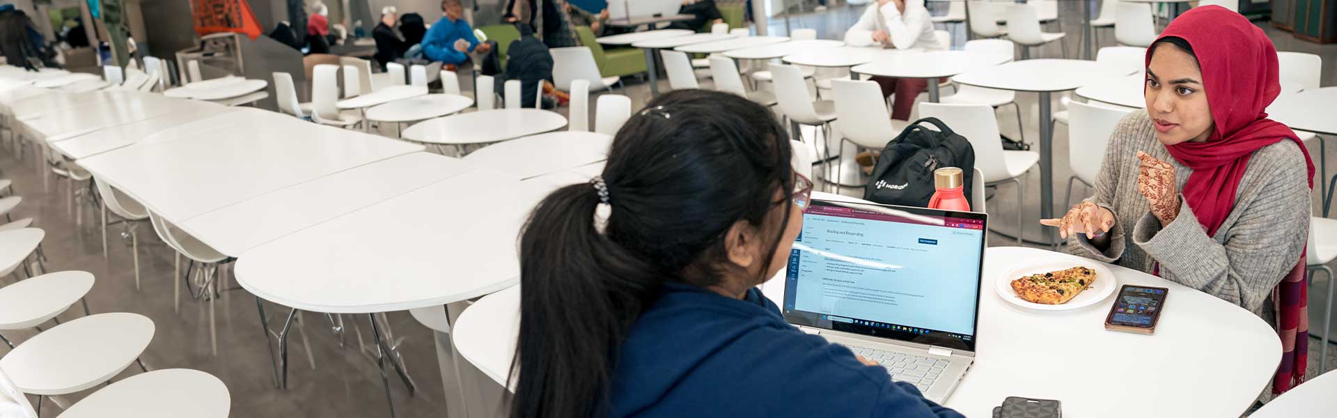 Two students sitting in across from each other in a cafeteria, talking and eating pizza