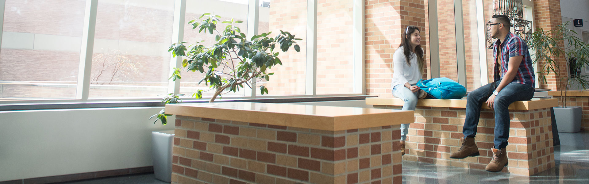 Two students conversing indoors while sitting on short brick wall