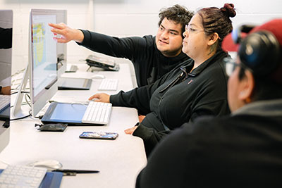 Three students discussing a project while pointing to a large computer screen
